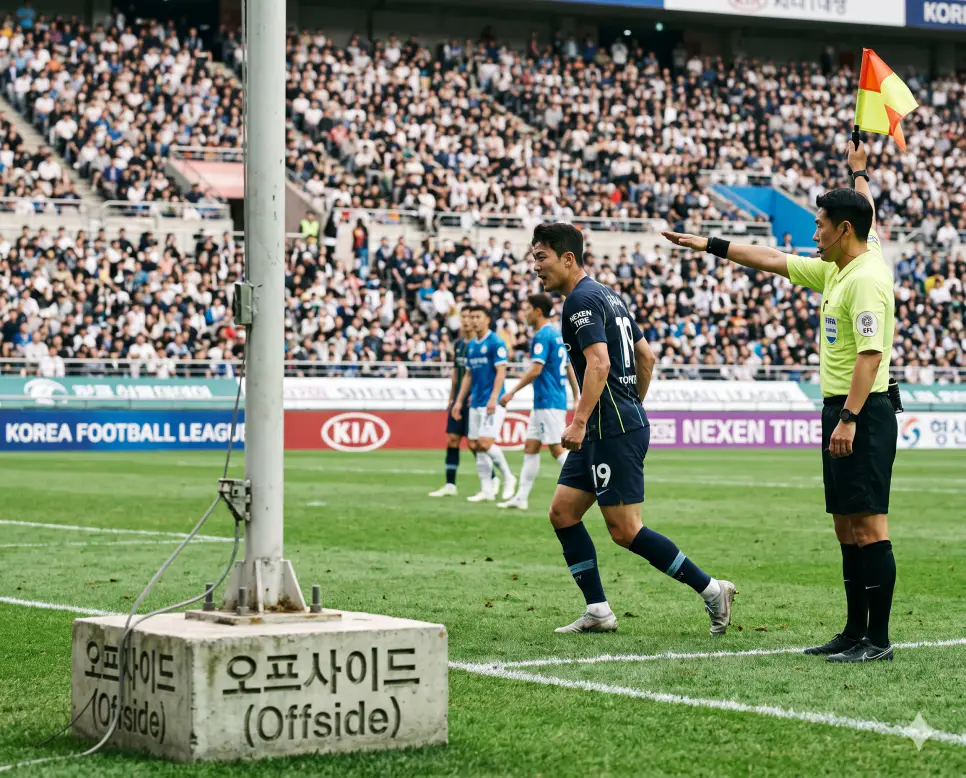 오프사이드 뜻, 세트피스 뜻, 킥오프 뜻: 축구 규칙 위반 정황과 정밀 전술 및 행정 명령의 본질 1 축구 경기에서 공격수가 상대 진영에서 패스가 유발되는 순간에 공과 상대의 두 번째 최후방 수비수보다 전방에 위치해 있을 때 선언되는 규칙 위반 정황인 오프사이드(Offside)의 실제 판정 순간을 축구 시즌 경기장 터치라인 부심이 정면을 향해 깃발을 수평으로 높이 들고 오프사이드 반칙을 선언하며 공격수가 허탈하게 멈춰 서는 뉴스 보도사진 스타일의 정면 극실사 이미지